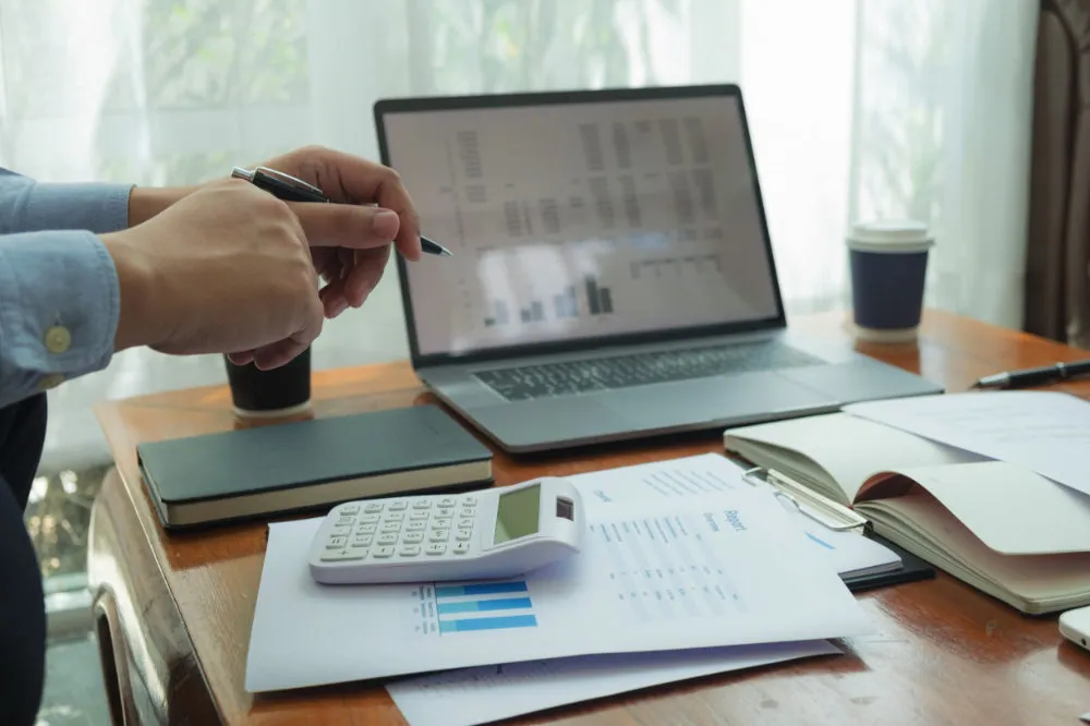 Person mit Stift an einem Holzschreibtisch, davor ein Laptop mit Finanzkennzahlen und Balkendiagrammen, ein Taschenrechner und ausgedruckte Berichte.
